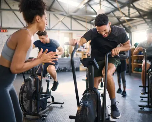 Bir adam kondisyon bisikletinde kardiyo yapıyor. Resmin solunda spor eğitmeni adama motivasyon veriyor. Arka planda başka bir adam kondisyon bisikletini kullanıyor