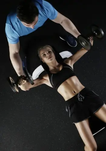 bir kadın dambıllarla bench press yapıyor, bir adam ona yardım ediyor